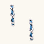 White gold-plated hoops intertwined with blue denim fabric in a twisted rope design on cream background.