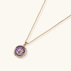 Gold-plated circular pendant with hand-painted purple enamel floral design on delicate chain against cream background