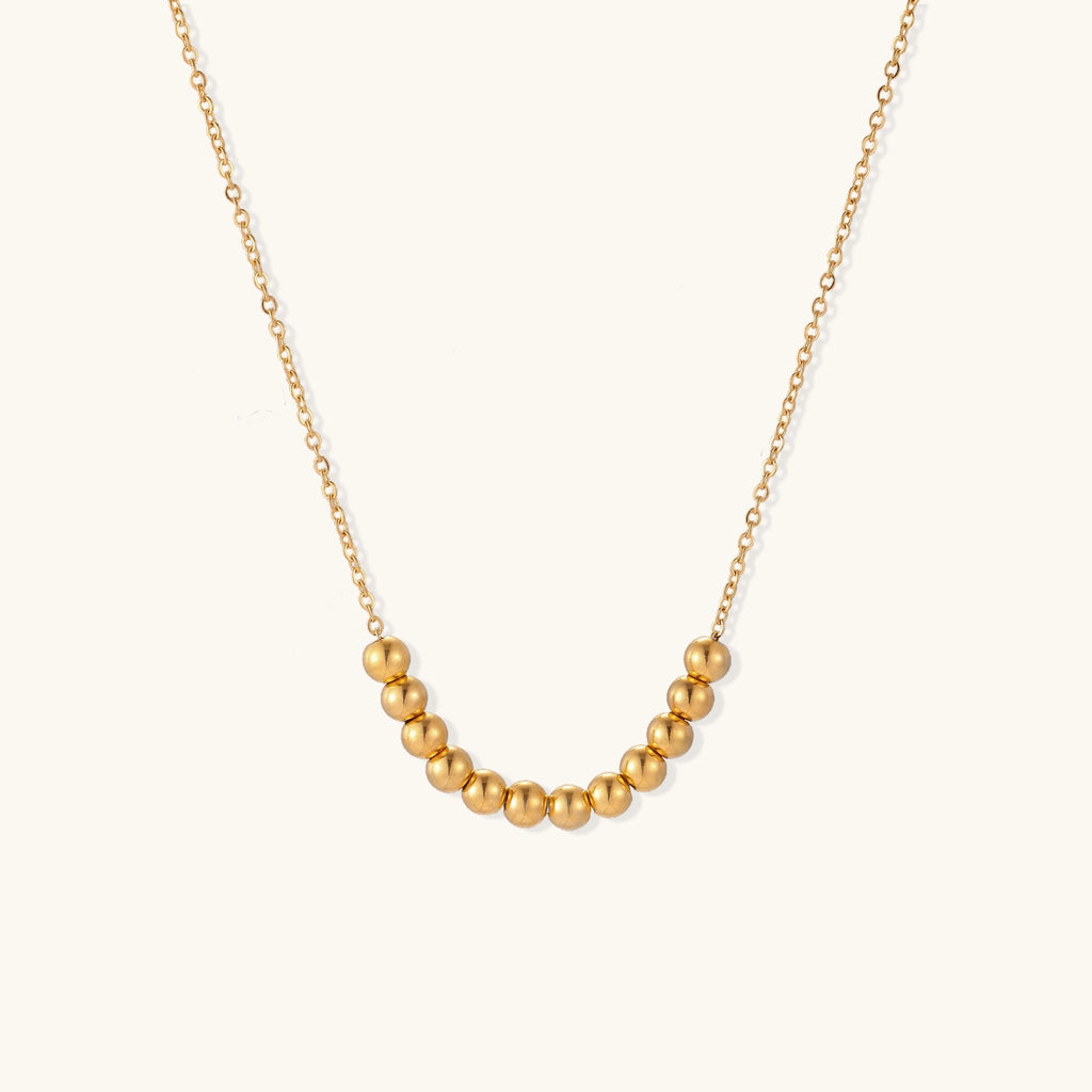 Delicate gold-plated necklace with graduated spherical beads forming gentle smile curve on beige background