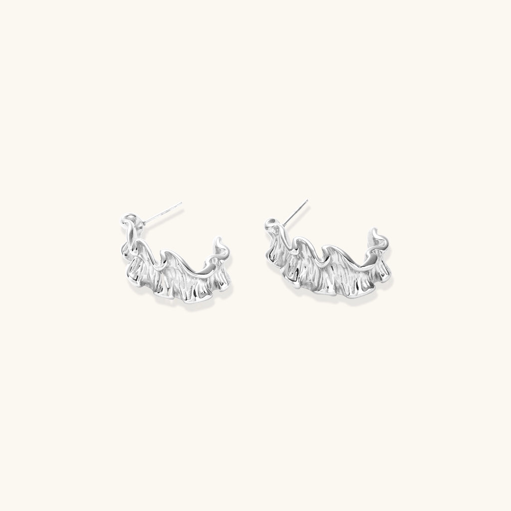 White gold plated sculptural hoop earrings with organic wavy edges and reflective metallic finish on cream background