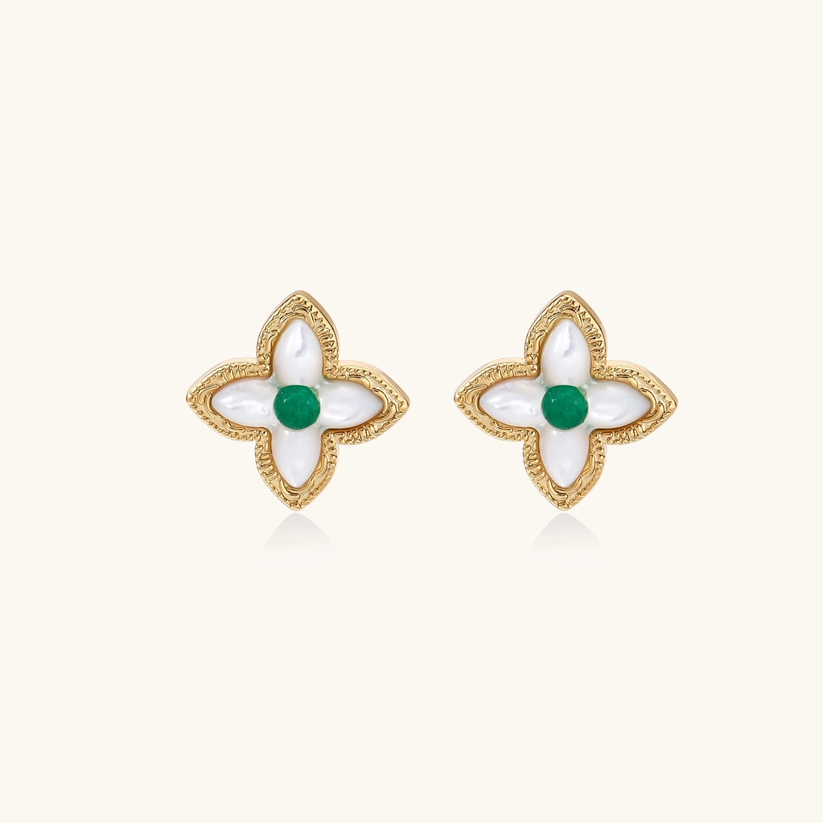 Gold quatrefoil stud earrings with white mother-of-pearl petals and emerald green gemstone center on beige background.